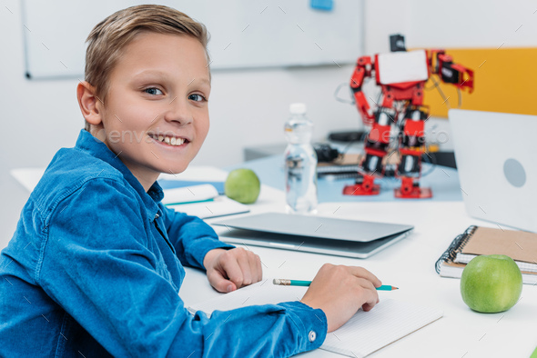 schoolboy sitting at desk with robot model, looking at camera and writing in notebook during STEM