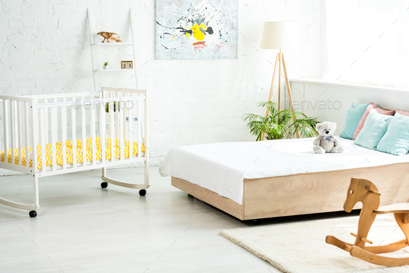 baby crib near bed with white bedding and pillows near teddy bear and rocking horse