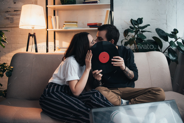beautiful young couple covering faces with vinyl record and kissing while sitting on couch in living
