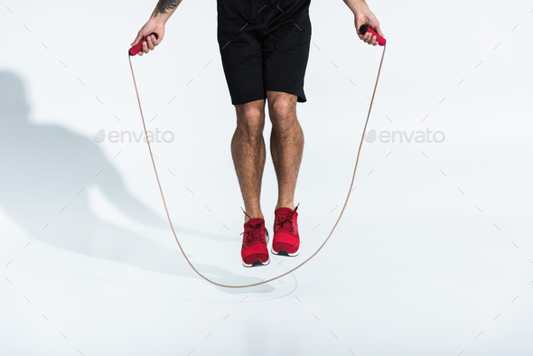 partial view of man in black shorts and red sneakers jumping with skipping rope on white