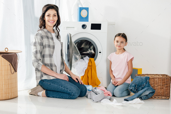 daughter in pink t-shirt and mother in grey shirt sitting on floor near scattered clothes and