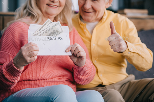 cropped view of senior couple showing thumb up sign while holding envelope with 'roth ira' lettering - Stock Photo - Images