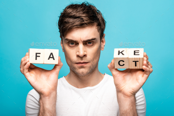 angry man showing wooden cubes with fake fact lettering and looking at camera isolated on blue