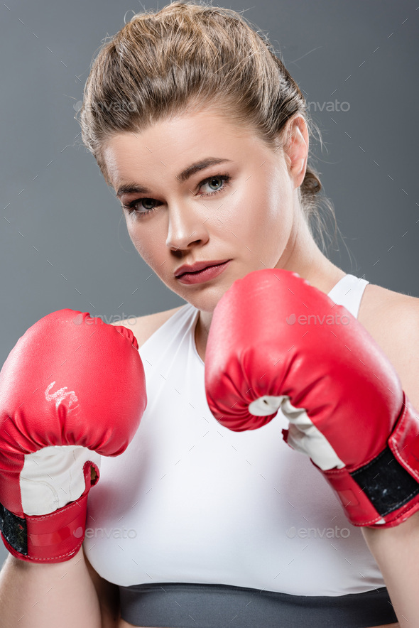 beautiful young woman in boxing gloves looking at camera isolated on grey
