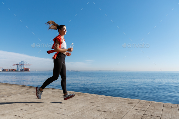 Side view of athletic young woman running along the seaside promenade. Sportswoman jogging near the
