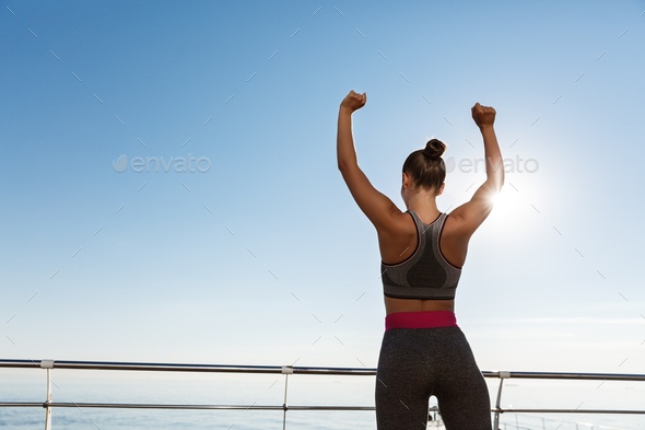 Rear view of happy and satisfied fitness woman looking at the sea and raising hands up like champion