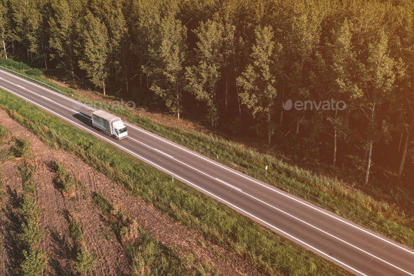 Aerial view of lorry truck on the road through green forest landscape in summer afternoon, drone pov - Stock Photo - Images