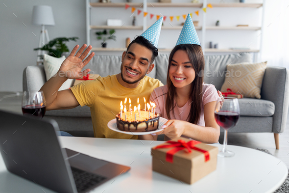 Online birthday celebration. Multiracial couple having video call, showing cake with candles at