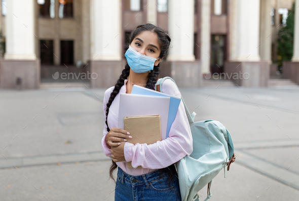 Indian female student in medical mask posing outdoors with notepad and books, standing near
