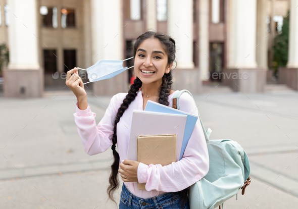 Happy indian student removing medical mask, posing outdoors with notepads near university building