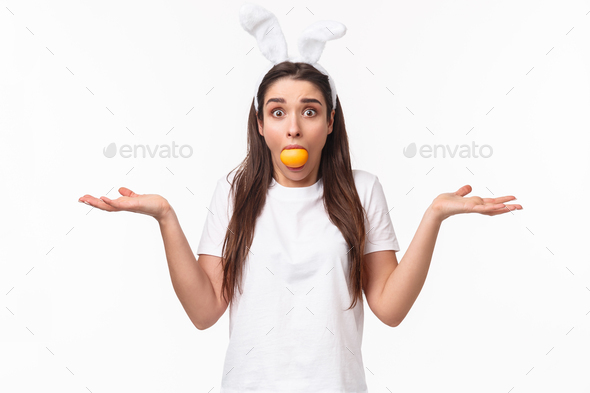 Portrait of confused and indeisive young funny woman with rabbit ears, holding colored Easter egg in