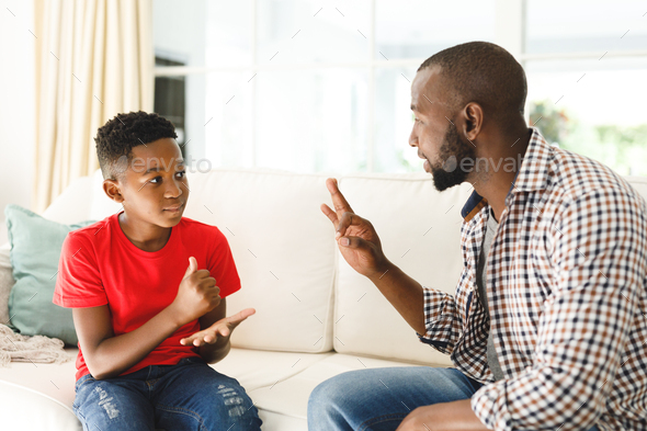 Happy african american father with son sitting on couch in living room talking sign language - Stock Photo - Images