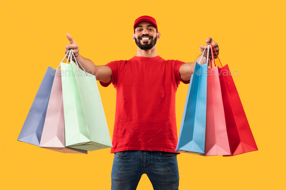 Middle-Eastern Male Courier Giving Shopper Bags Delivering Clothes, Yellow Background