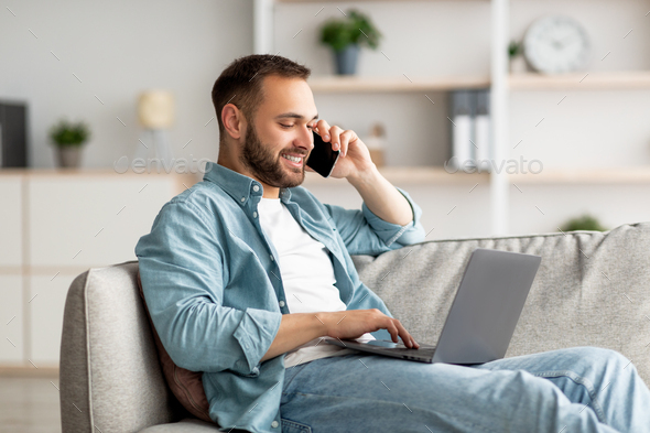 Attractive young guy talking on smartphone and using laptop computer on couch at home office - Stock Photo - Images