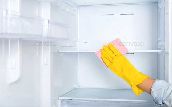 A woman\'s hand in a yellow rubber protective glove and sponge washes and cleans refrigerator shelves