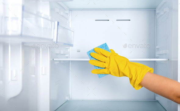 A woman\'s hand in a yellow rubber protective glove and a blue sponge cleans the refrigerator shelves
