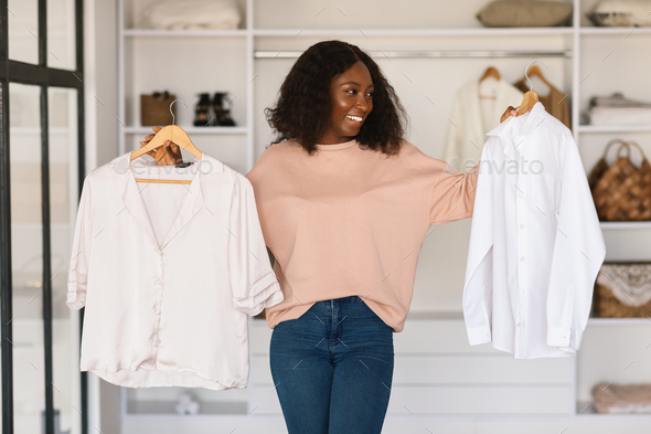 Happy African Woman Shopping And Choosing Clothes Holding Shirts Indoors - Stock Photo - Images