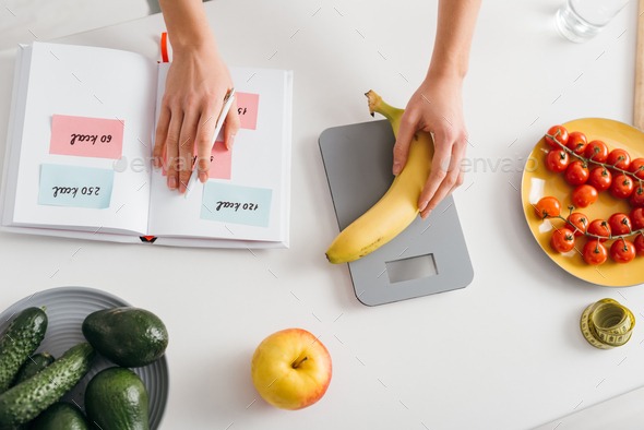 Top view of girl putting banana on scales while writing calories in notebook, calorie counting diet