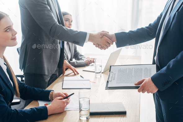 Side view of employee with resume shaking hands with recruiter during job interview in office