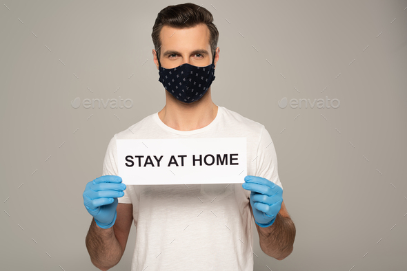 Man in safety mask and latex gloves holding card with stay at home lettering isolated on grey