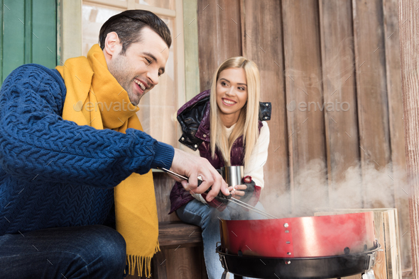 Smiling young woman with hot drink looking at happy man grilling meat on porch