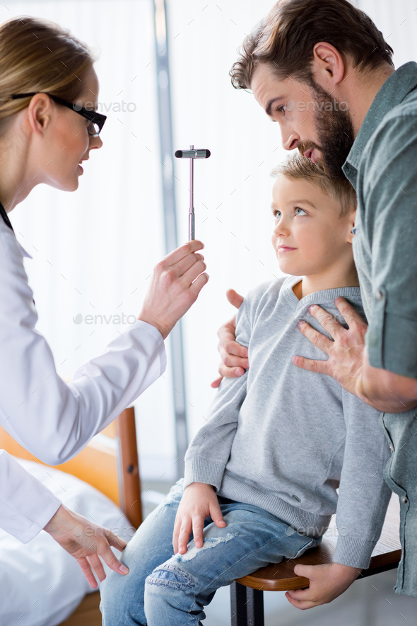 Female doctor with reflex hammer inspecting little boy sitting near father