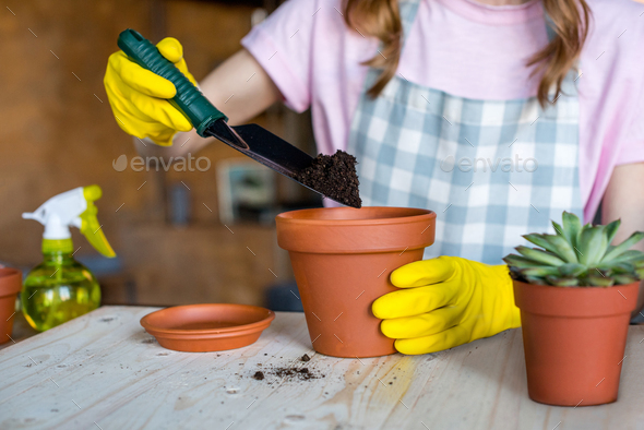 young woman in rubber gloves heaping ground in flowerpot with garden spade