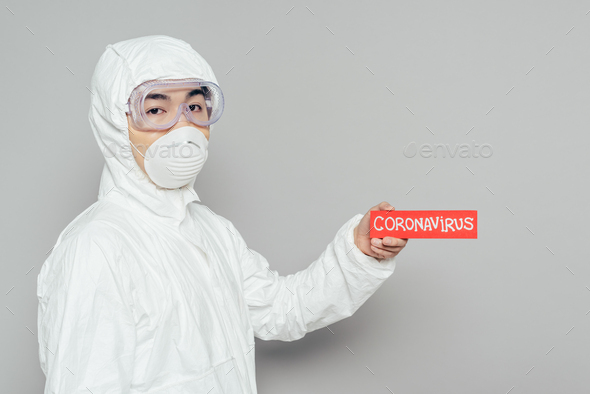 asian epidemiologist in hazmat suit and respirator mask holding warning card with coronavirus