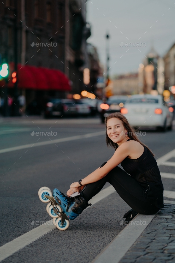 Woman poses on asphalt puts on roller-skates being in good mood spends free time riding rollerblades