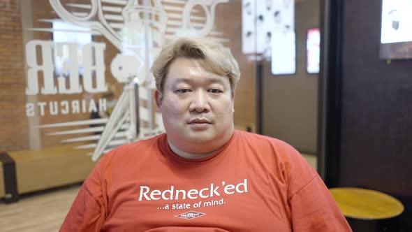A Barber Wearing Orange Tshirt Sitting in His Shop and Being Videoed By a Camera alt