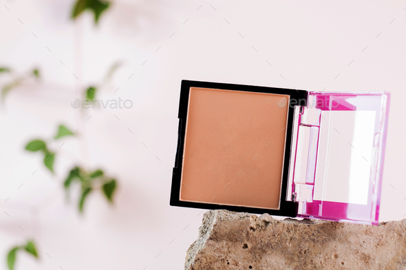 Open case with powder on grey stone on light pink background with blurred plant