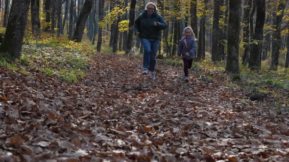 A woman and child run through the fallen leaves in the woods. In the background is a young dog alt