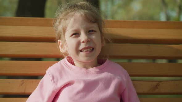 Joyful Girl Without Tooth Sits on Bench in Spring Park alt