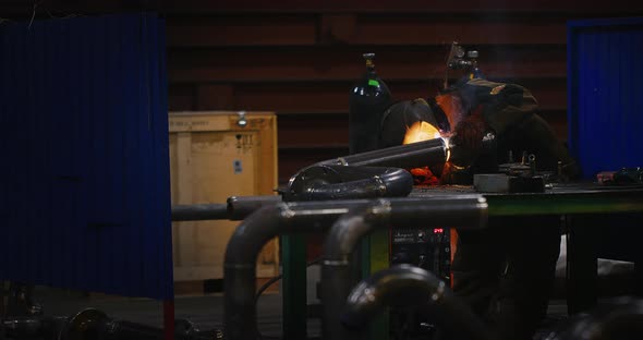 Welder in a Welding Helmet in Work Clothes Welds a Pipe at the Factory alt