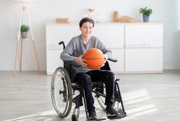 Happy disabled teenage boy holding basketball, sitting in wheelchair and smiling at camera indoors - Stock Photo - Images