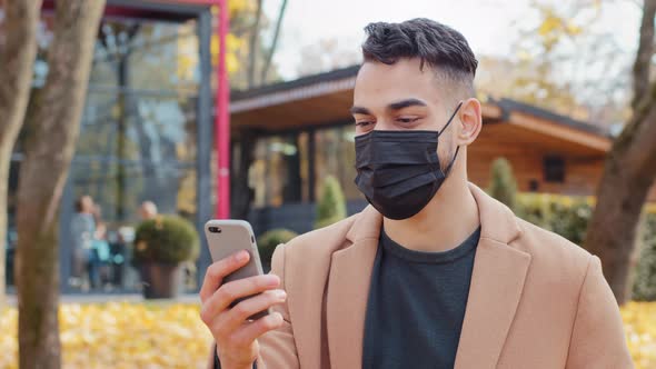 Spanish Young Man in Medical Mask Talking on Video Call Using Smartphone Standing in Park Hispanic alt