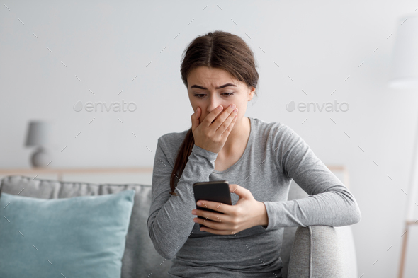 Shocked sad despaired caucasian millennial female covering her mouth after reading bad news in phone