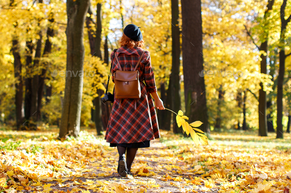 redhead girl walks through the park hold dry yellow fallen leaf at sunny day, back view. Autumn mood - Stock Photo - Images