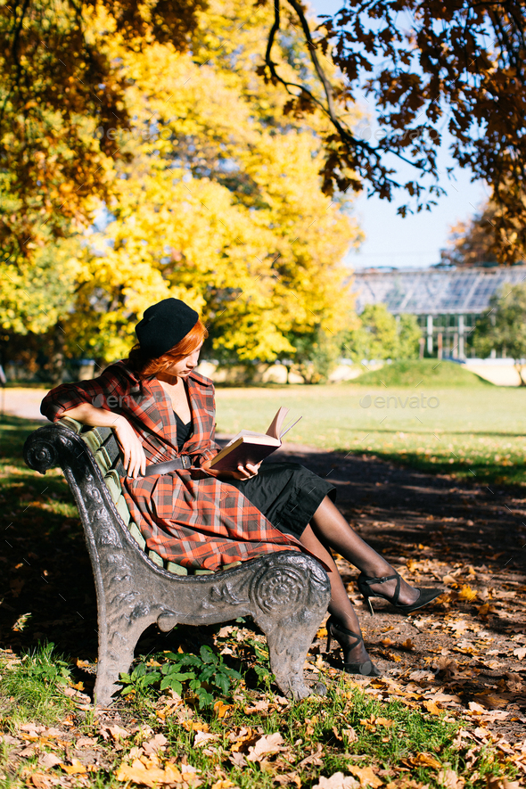redhead young woman in plaid coat and black beret reading book on bench in autumn park at sunny day - Stock Photo - Images