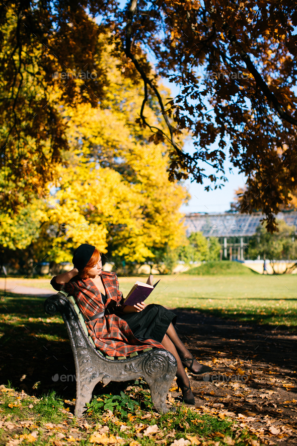 redhead woman in plaid coat and black beret reading book on bench in autumn park at sunny day - Stock Photo - Images