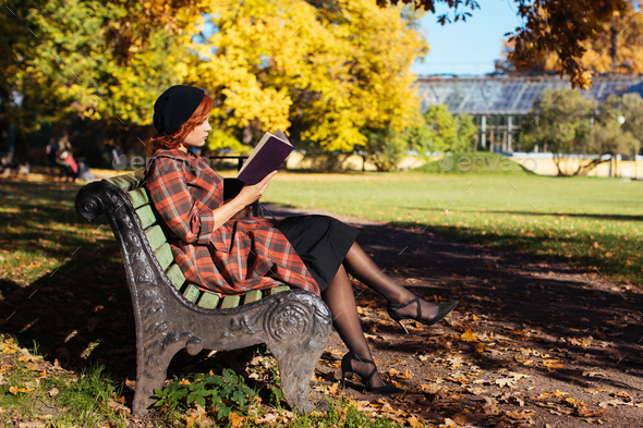 redhead woman reading a book on a bench in autumn park at sunny day. Autumn mood, leisure time - Stock Photo - Images