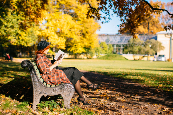 redhead girl in checked coat and beret reading book on bench, resting in autumn park at sunny day - Stock Photo - Images