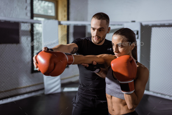 young male trainer holding hand of beautiful sportswoman in boxing gloves training in sport club