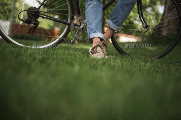 Lower section of man in jeans standing with bicycle on green grass