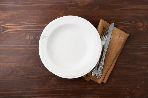 Empty white circle ceramic plate on wooden background with silverware and linen napkin