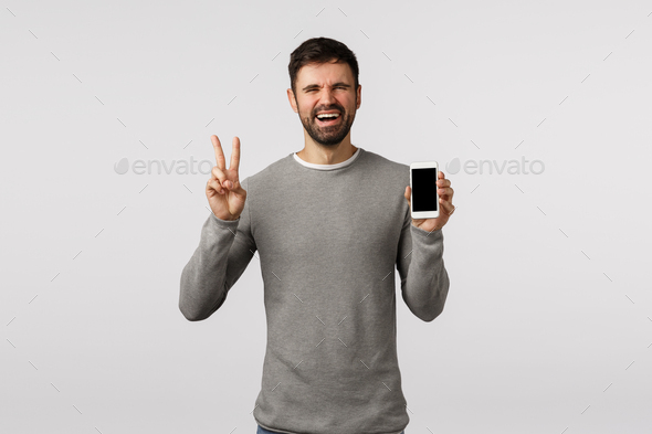 Funny and emotive, charismatic bearded man holding smartphone, make peace, victory sign, showing