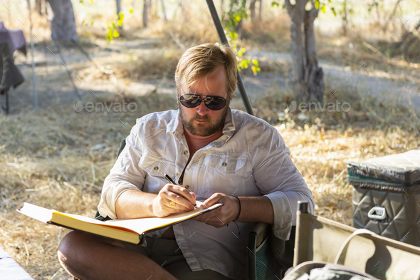 Bearded man wearing sunglasses sitting on chair in a safari camp, writing a journal