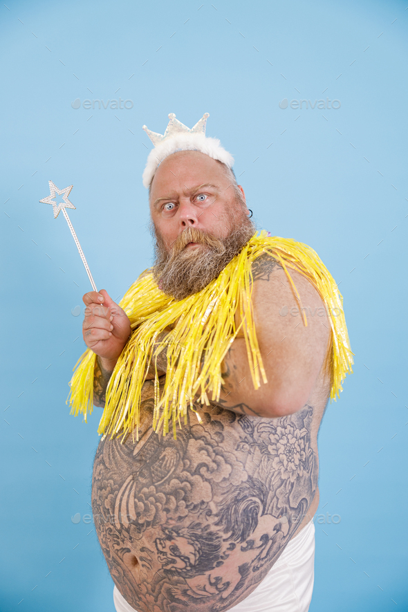 Doubting plus size man with crown and magic stick poses on light blue background