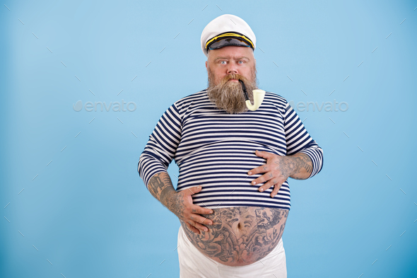 Middle aged obese man in sailor costume holds hands on tummy on light blue background