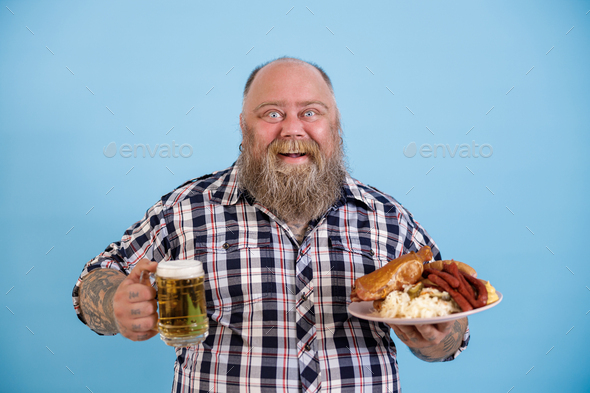 Bearded man with overweight holds greasy food and beer on light blue background